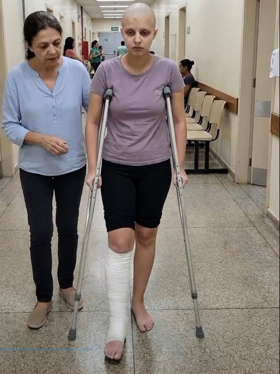 Marcela, adolescente com a perna imobilizada, caminhando de muletas ao lado da mãe no corredor do hospital